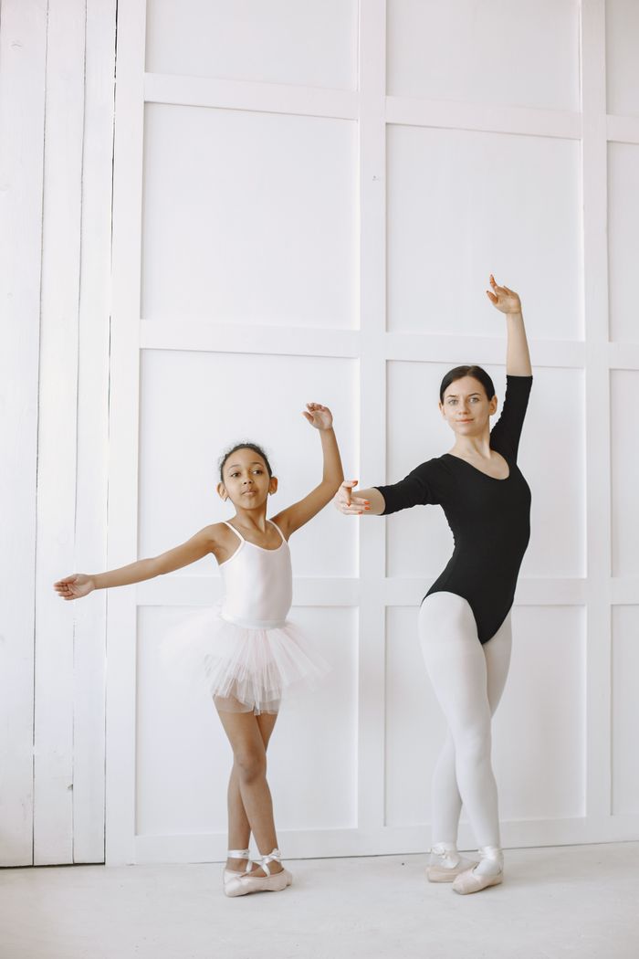 A young ballerina learning dance poses from her mentor in a bright indoor studio setting.